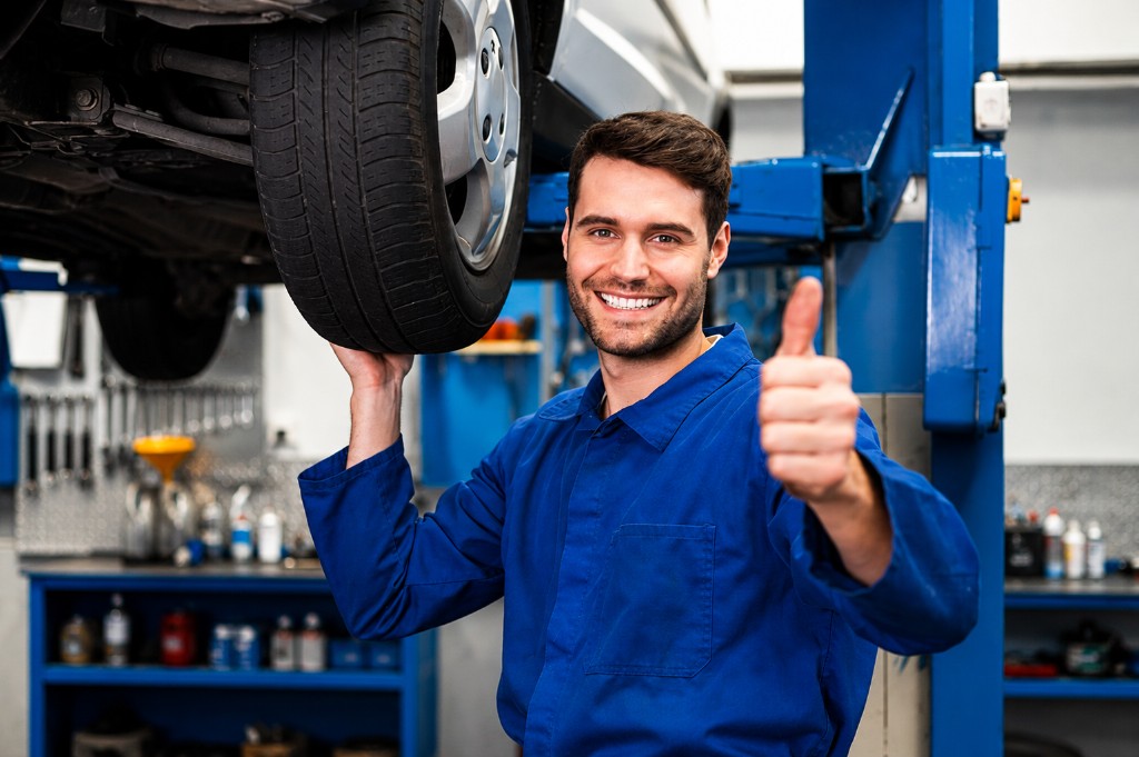 Professional automotive mechanic giving thumbs up in a workshop with a vehicle on a hydraulic lift